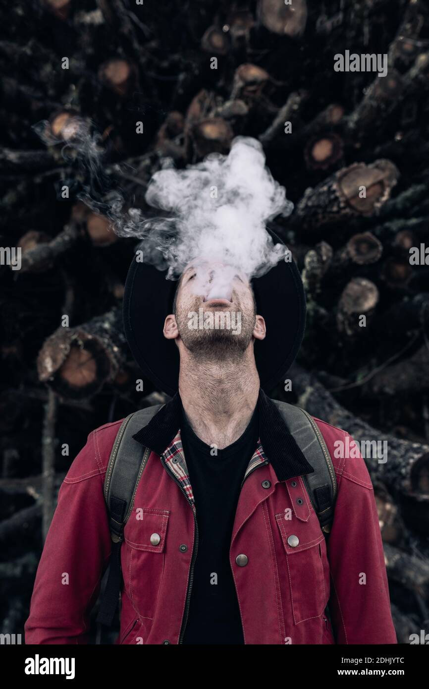 Hipster maschile in cappello e camicia rossa che esala nuvola di fumo in piedi con gli occhi chiusi contro pila di tronchi in natura Foto Stock