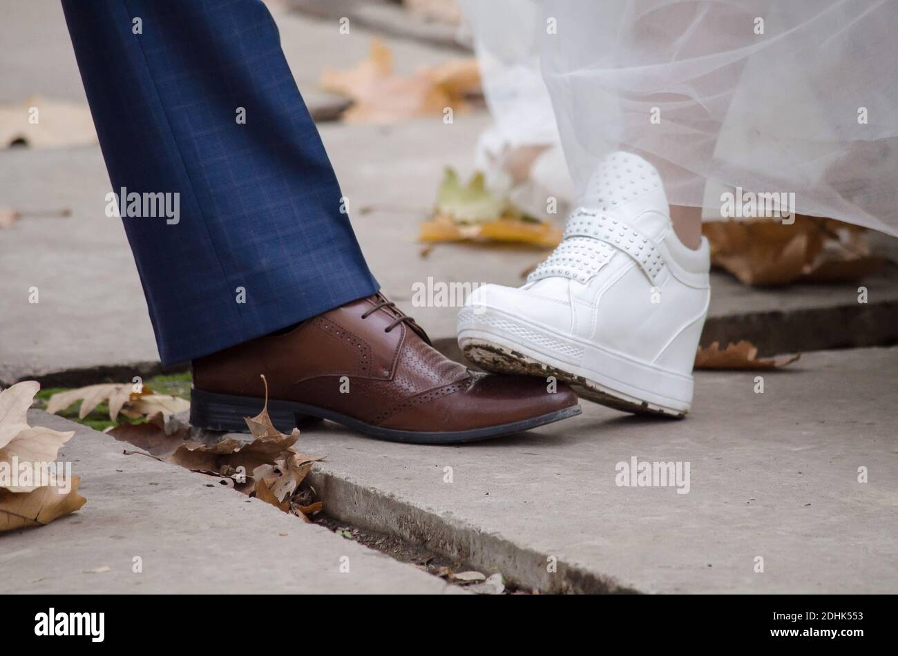Il piede della sposa in sneakers bianche calca il piede dello sposo su scarpe marroni, in autunno tra le foglie, primo piano Foto Stock