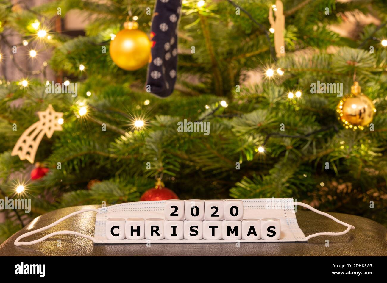 Simbolo del periodo natalizio durante la pandemia della corona. I dadi formano l'espressione 'Natale 2020' sulla parte superiore di una maschera di fronte ad un albero di Natale Foto Stock