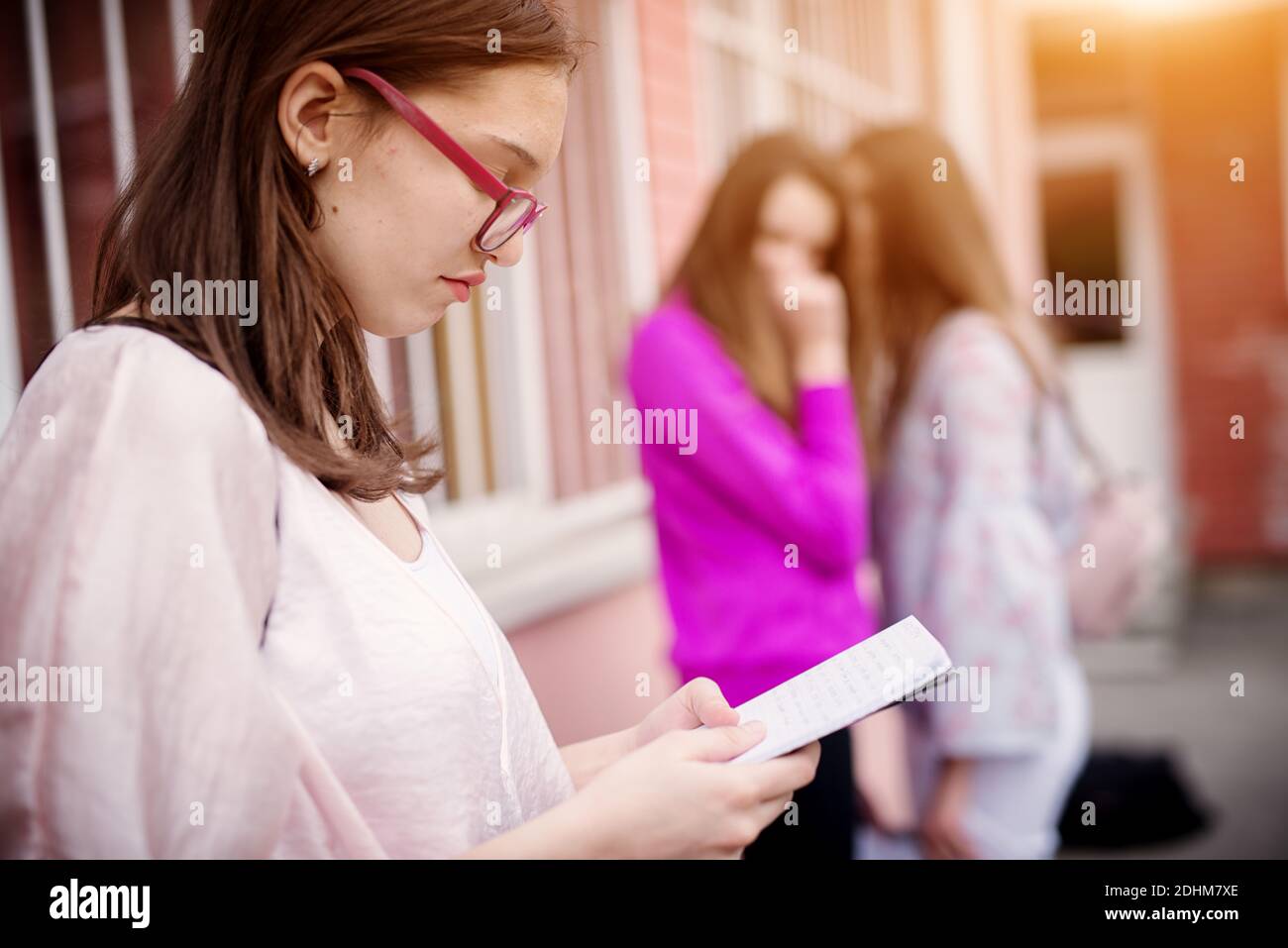 Triste infelice ragazza della scuola superiore che legge dalle note della scuola mentre i suoi compagni di scuola la pettegolano davanti mentre si levano in piedi fuori della scuola. Foto Stock