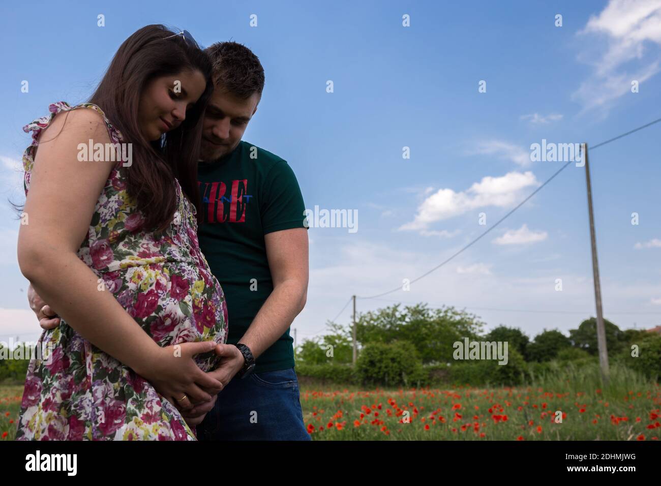 Giovane coppia con donna incinta in piedi su un prato pieno Dei fiori comuni di papavero Papaver Rhoeas e guardandola urto del bambino Foto Stock