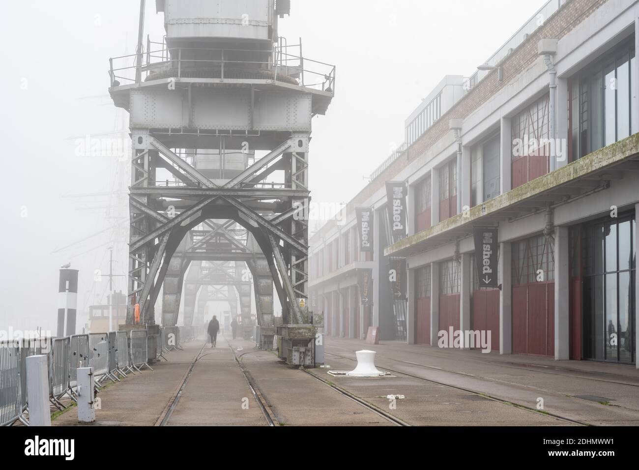 I pedoni camminano attraverso la nebbia passando davanti al museo M Shed e alle storiche gru portuali sul porto galleggiante di Bristol. Foto Stock