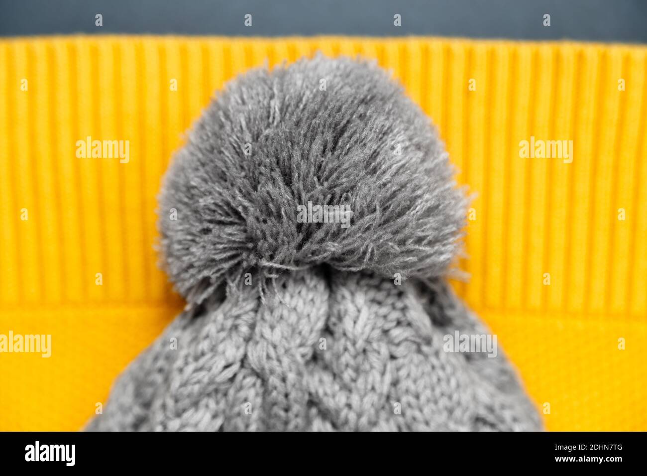 Primo piano del cappello grigio lavorato a maglia con pompon sul pullover giallo nei colori alla moda su sfondo grigio. Colori dell'anno 2021. Abiti a maglia, caldi Foto Stock