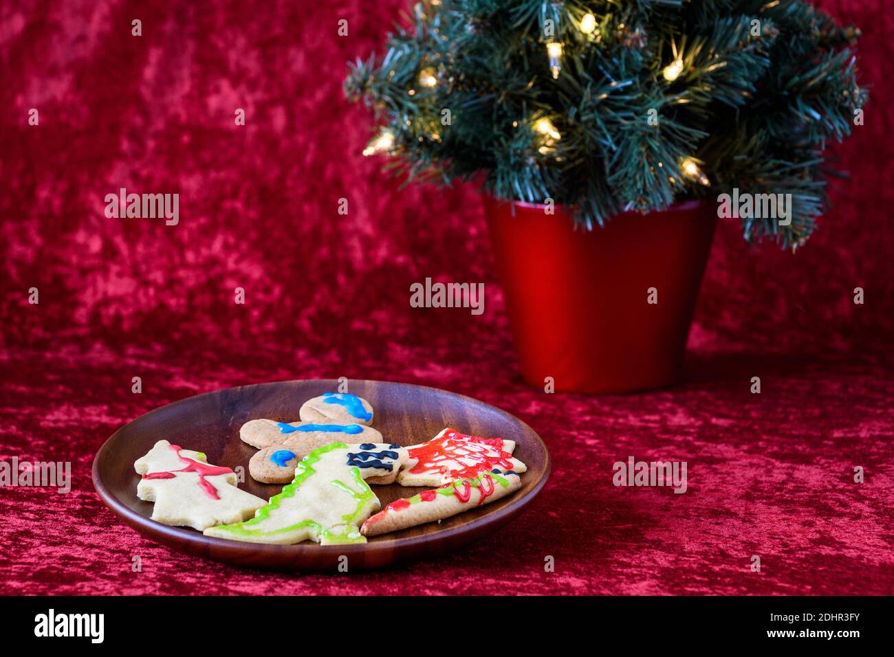 Biscotti allo zucchero appena sfornati decorati dai bambini per le delizie natalizie, su un piatto di legno, con albero illuminato, su sfondo rosso Foto Stock