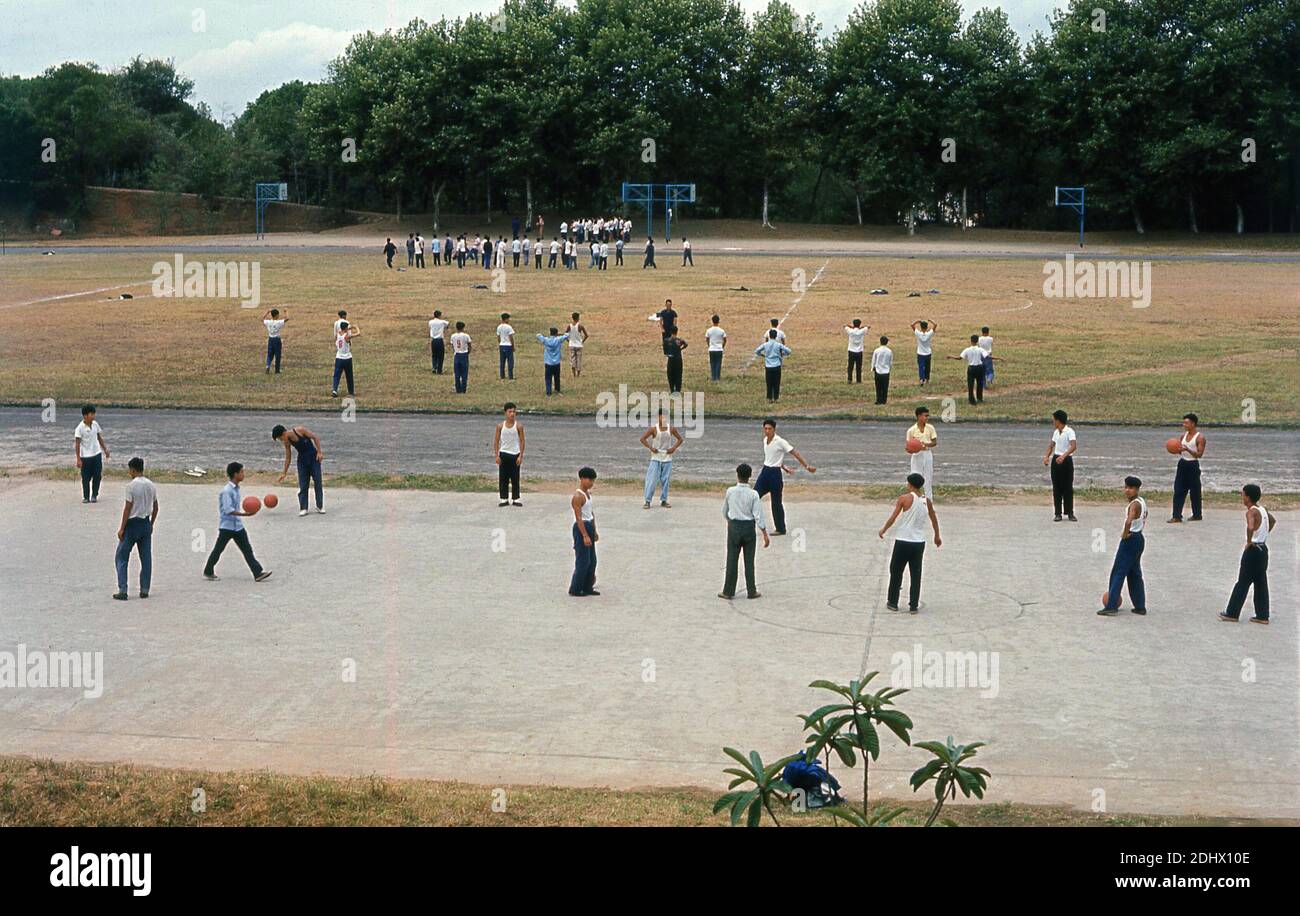 Anni '60, storico, Wuhan, Cina, gruppi di studenti di scuola superiore maschile o universitari fuori giocare con un basket su una zona ghiaia e fare esercizi su un campo sportivo. Gli esercizi di gruppo sono stati una parte importante della vita cinese in quest'epoca, un periodo comunemente noto come 'Cina maoista', in quanto il paese è stato guidato per molti anni dal presidente Mao Zedong che ha introdotto quella che è chiamata la rivoluzione culturale Foto Stock