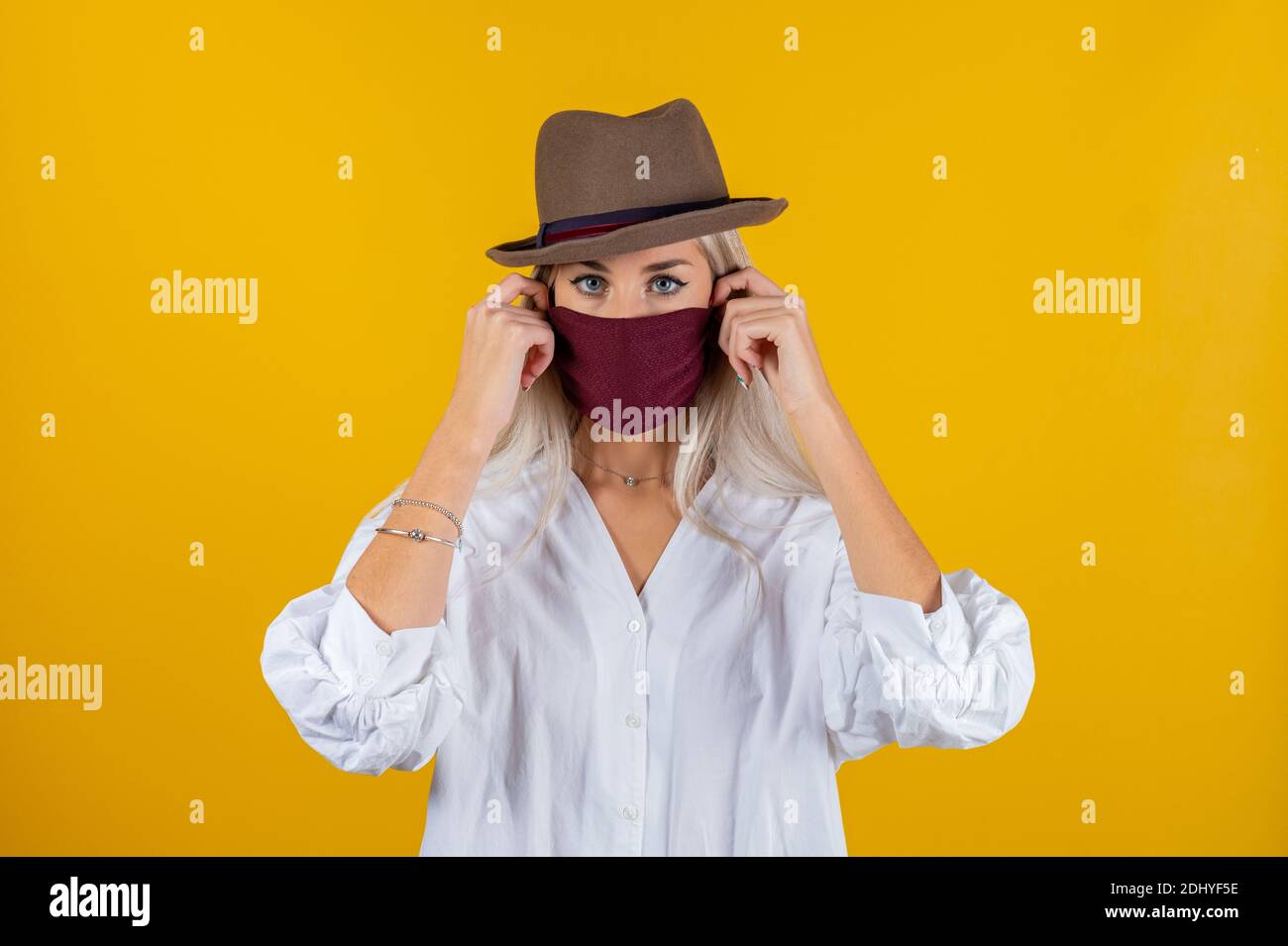 Donna che indossa un'elegante maschera protettiva per il viso e un cappuccio marrone, che si posa su sfondo giallo. Accessorio moda alla moda durante la quarantena di Coronavirus pande Foto Stock