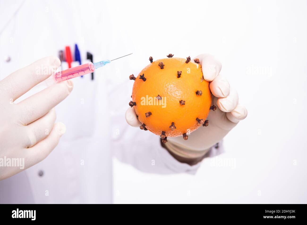 Mani del medico, guanti di vit coperti, tenendo una vaccinazione (siringa) e una paricola del virus, che è un arancione con chiodi di garofano Foto Stock