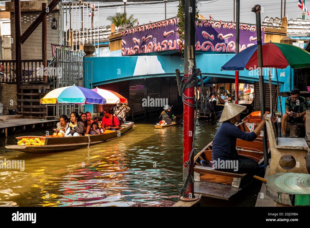 Una donna in una piccola san Pan offre spuntini per la vendita ai turisti in barca più grande con ombrelloni colorati al mercato galleggiante Damneon. Foto Stock Una donna in una piccola san Pan offre spuntini per la vendita ai turisti in barca più grande con ombrelloni colorati al mercato galleggiante Damneon. Foto Stock