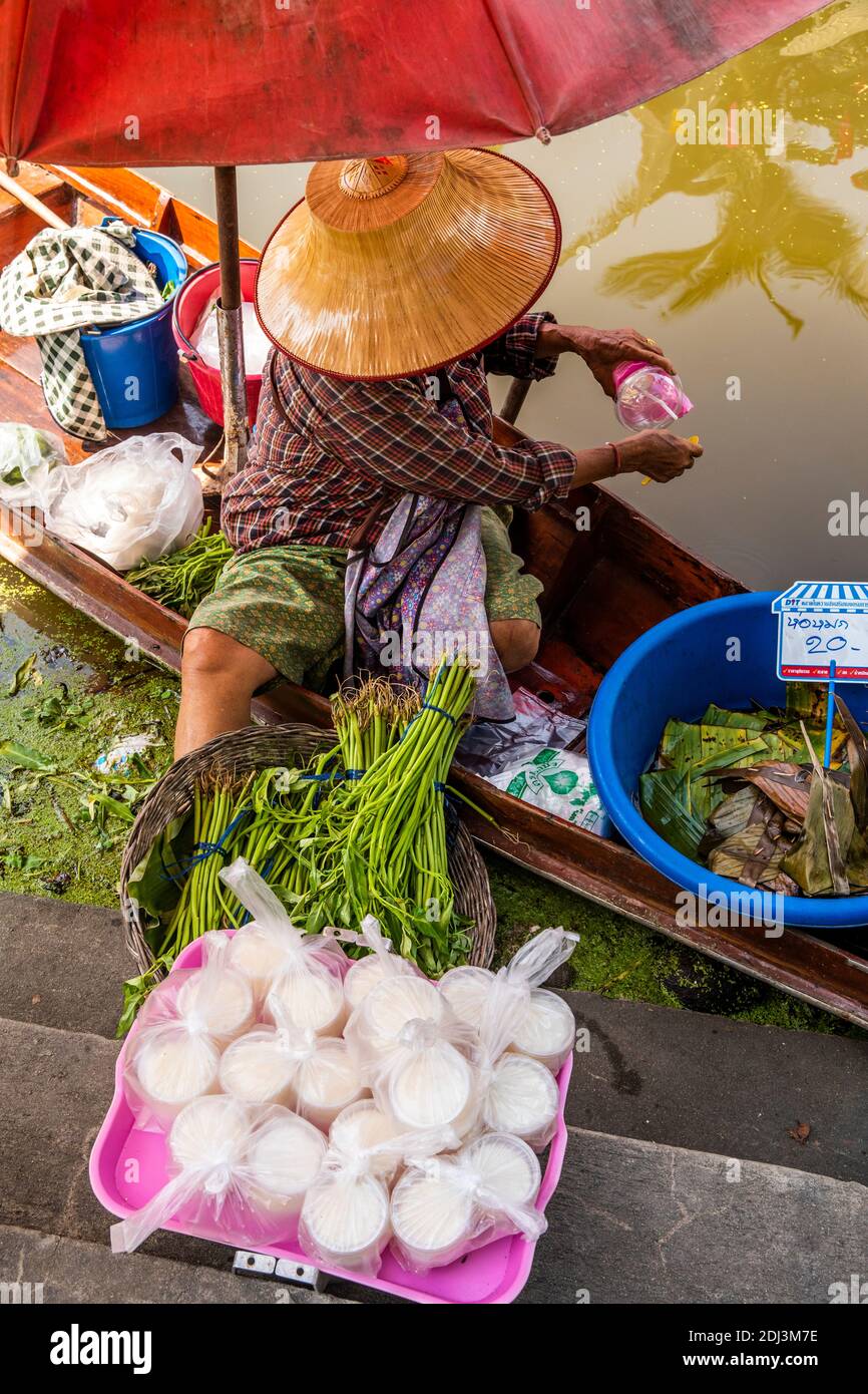 Sotto la protezione del suo cappello thailandese e un ombrello, una donna in. Il mercato di Amphawa lavora sulla sua barca da cui vende prodotti e riso. Foto Stock