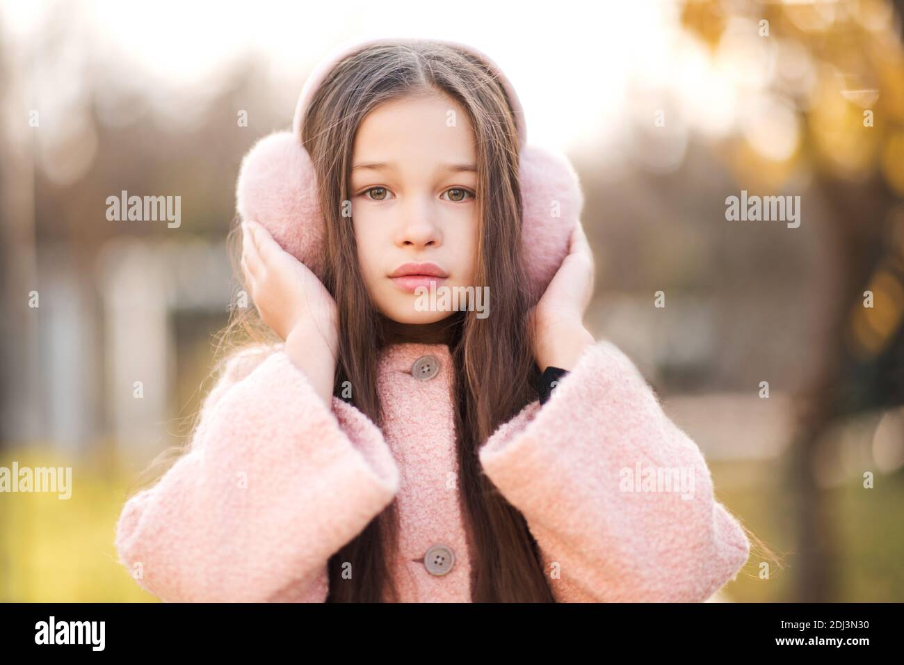 Bella ragazzina di 5-6 anni con cuffie rosa soffici e cappotto elegante su sfondo natura da vicino. Guardando la fotocamera. Infanzia Foto Stock