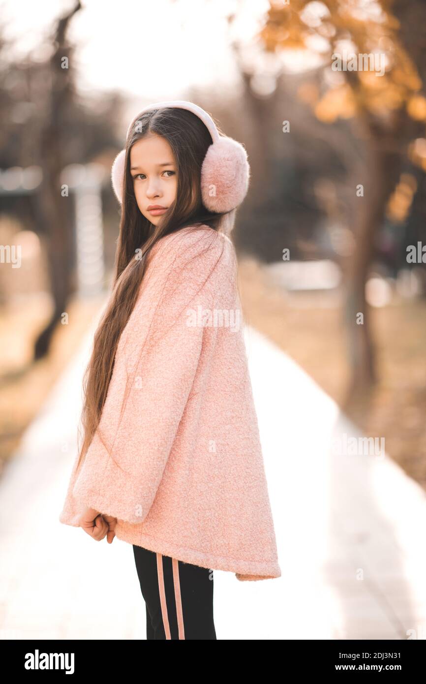 Elegante bambina di 5-6 anni con cuffie rosa soffici e giacca in piedi sulla strada della città in parco su sfondo natura all'aperto. Guardando Foto Stock