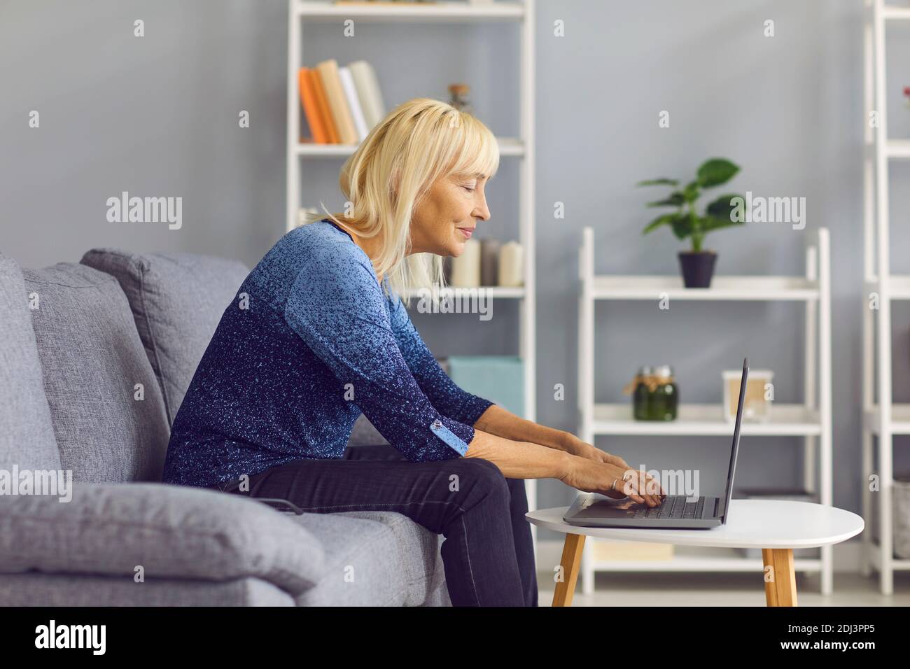 Vista laterale di una donna matura che scrive sul computer portatile mentre si siede sul divano a casa. Foto Stock