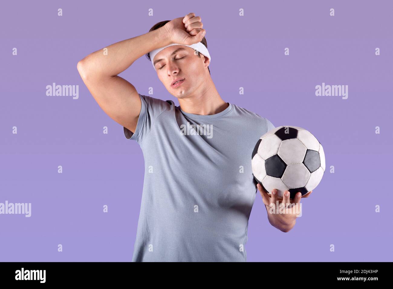 Giocatore di calcio stanco con palla che strofina sudore dopo la partita o. campionato su sfondo lilla studio Foto Stock