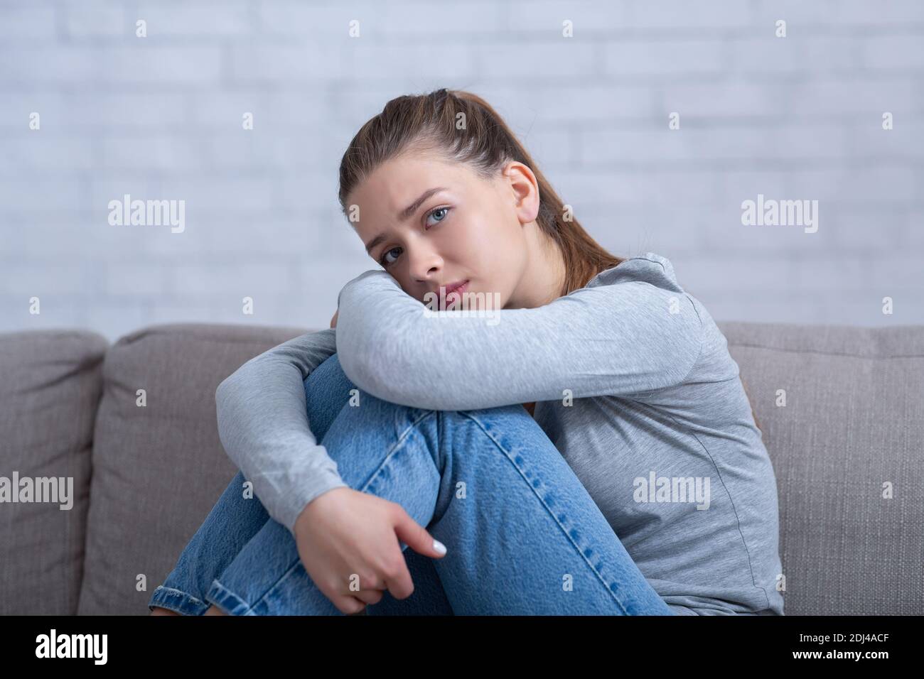 Giovane donna depressa che le aggira le ginocchia sul divano a casa, assorbita nei pensieri negativi, avendo disturbi mentali Foto Stock