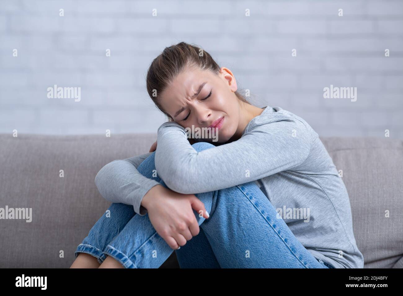 Giovane donna depressa seduta sul divano con gli occhi chiusi e abbracciando le ginocchia, sentendosi disperata e stressata Foto Stock