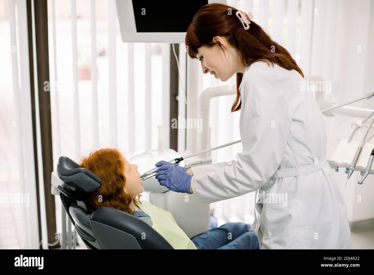 Odontoiatria per bambini, odontoiatria pediatrica. La giovane stomatologa caucasica femmina sta trattando i denti di una ragazza dai capelli rossi piuttosto in età scolare, usando il dente Foto Stock