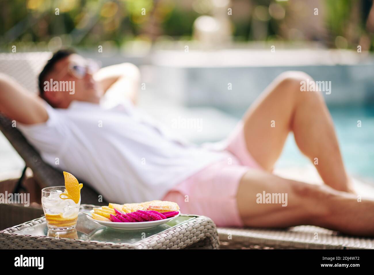Piatto con frutta tagliata e bicchieri di gin e tonico con la pelatura di gamma sul tavolo accanto all'uomo che poggia sopra chaise-lounge Foto Stock