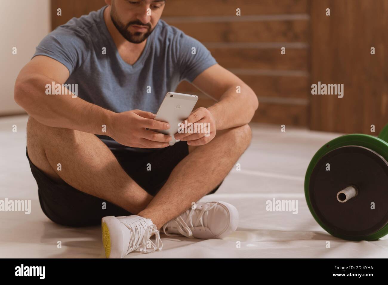 Body shot di bel giovane uomo alla ricerca online o testing seduta sul pavimento con smartphone, tono nero e verde barbell fitness, attrezzature per Foto Stock