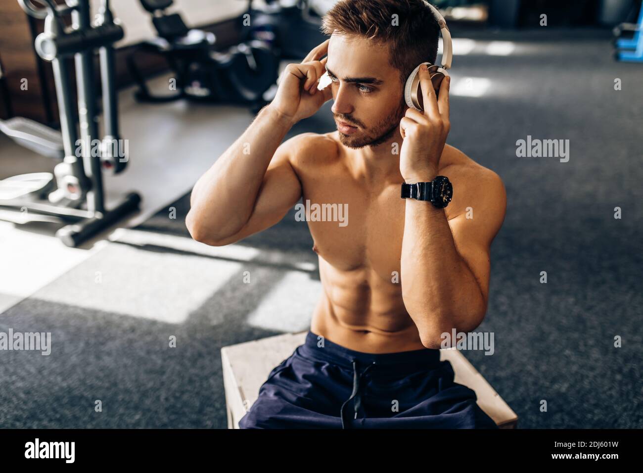 Giovane uomo in forma muscolare, irreprensibile, rilassante dopo l'allenamento, ascoltando musica in cuffia wireless, in palestra Foto Stock