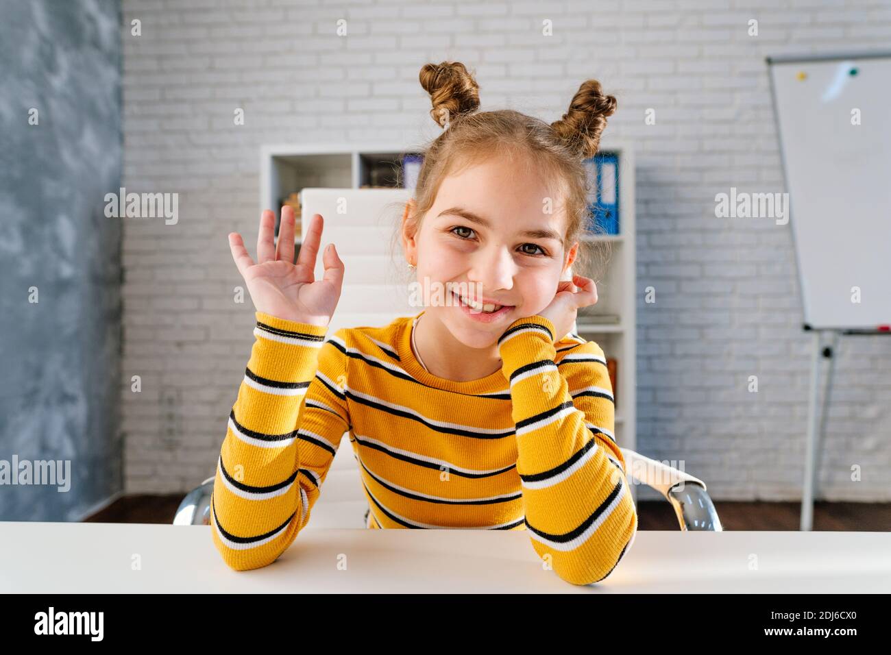 Felice sorridente 8 anni ragazza dire ciao guardando la macchina fotografica seduta alla scrivania nella stanza dei bambini durante la lezione online. Foto Stock