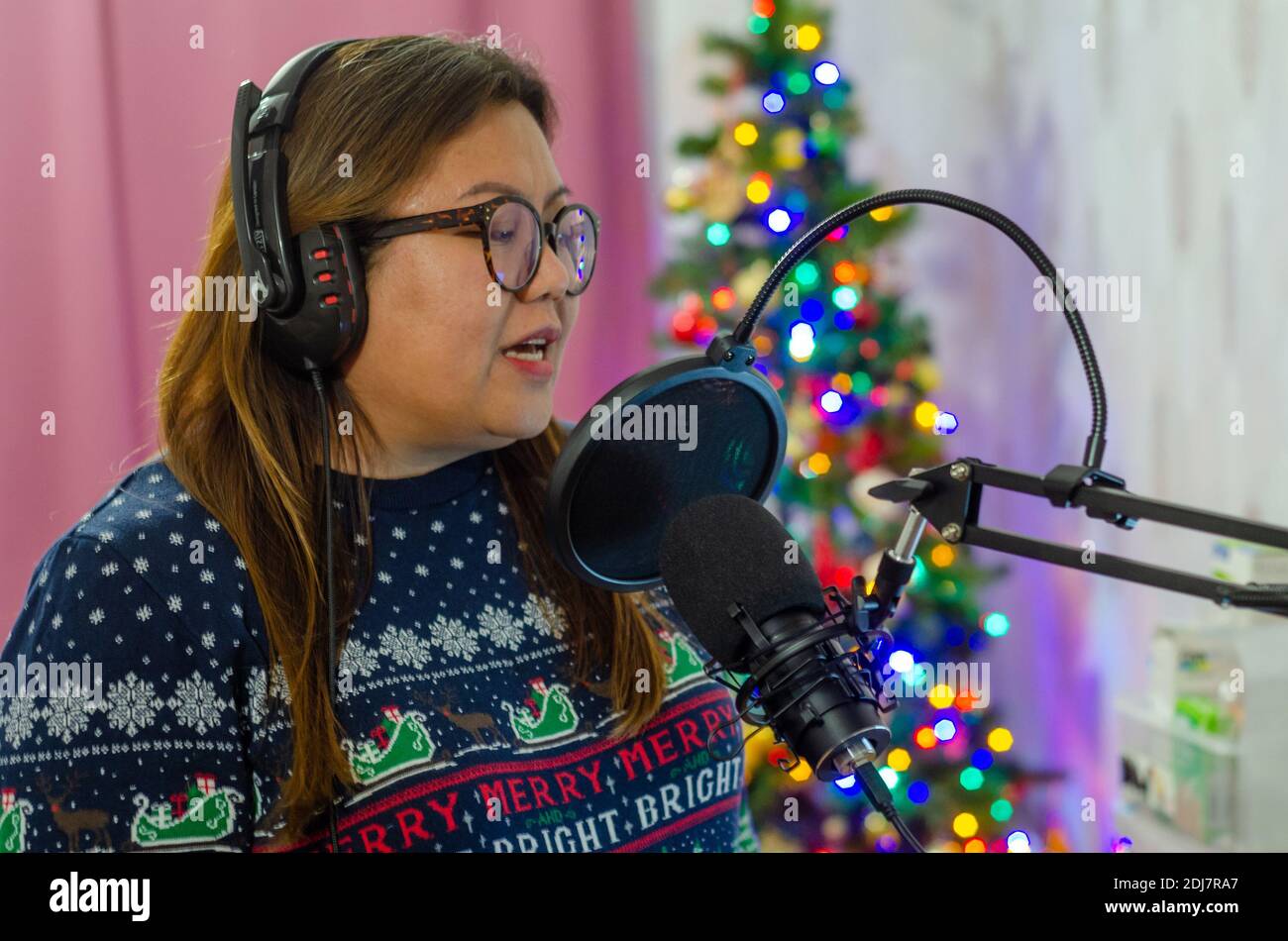 Una donna che indossa un jumper natalizio e indossa un set di cuffie che cantano in un microfono, registrando voci per una canzone. Foto Stock