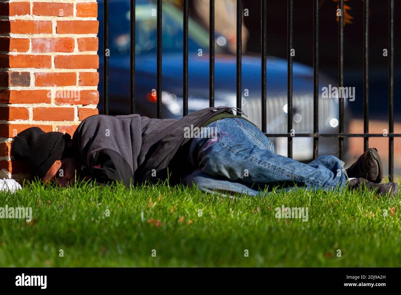 Alexandria, VA, USA 11-28-2020: Un uomo latino senza casa che indossa felpe e jeans sta dormendo sull'erba all'aperto in un parco cittadino da un muro di mattoni a. Foto Stock