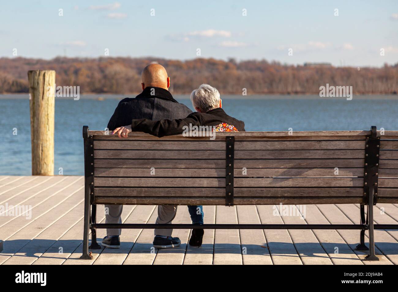 Una donna caucasica anziana con capelli grigi corti e un giovane afroamericano con la testa rasata sono seduti su una panchina su un molo che guarda al mare. IO Foto Stock