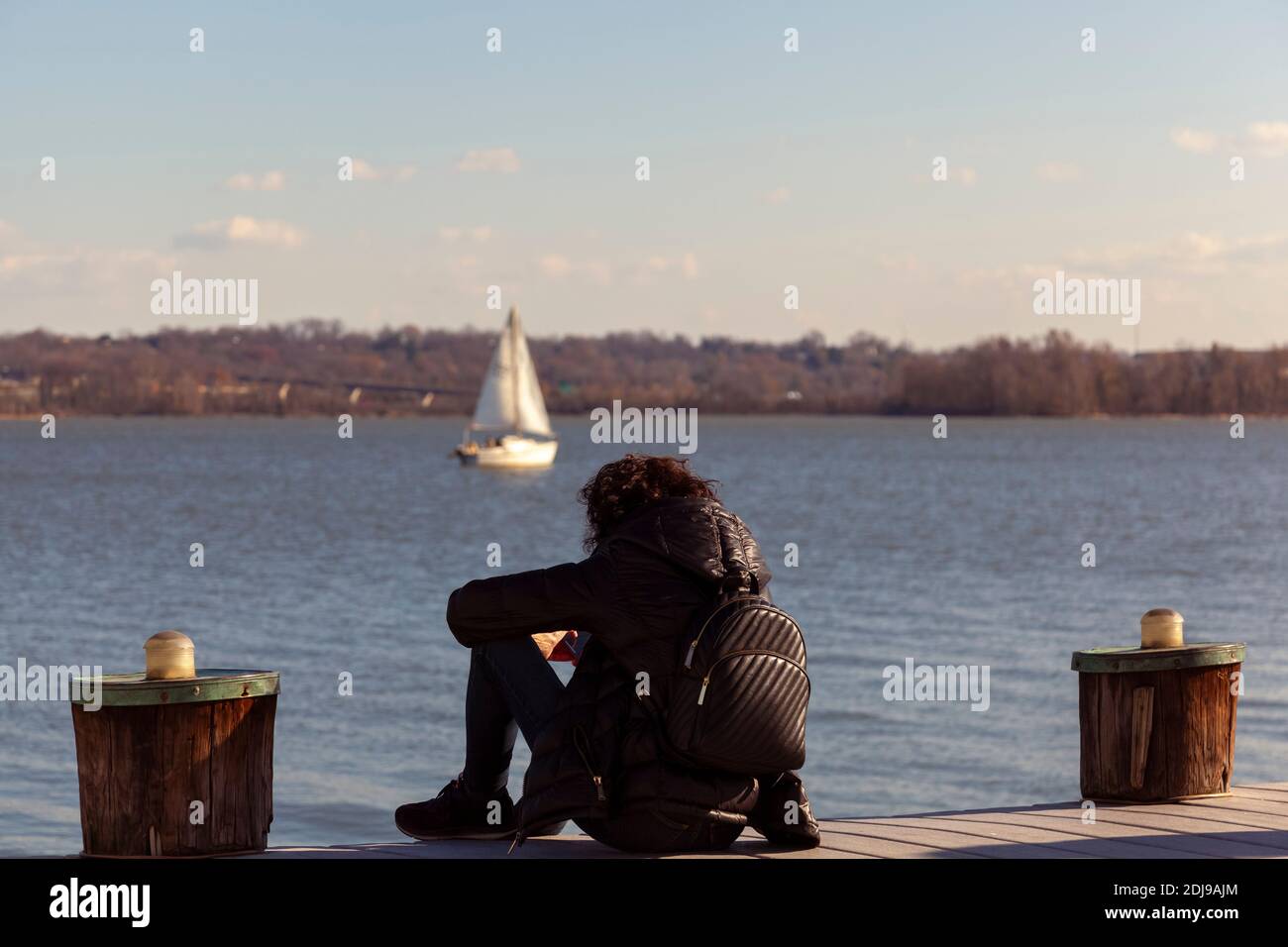 Una donna che indossa abiti scuri tra cui sneakers, cappotto invernale e zaino è seduta da sola su un molo che guarda al mare. Immagine per umore giù, premere Foto Stock