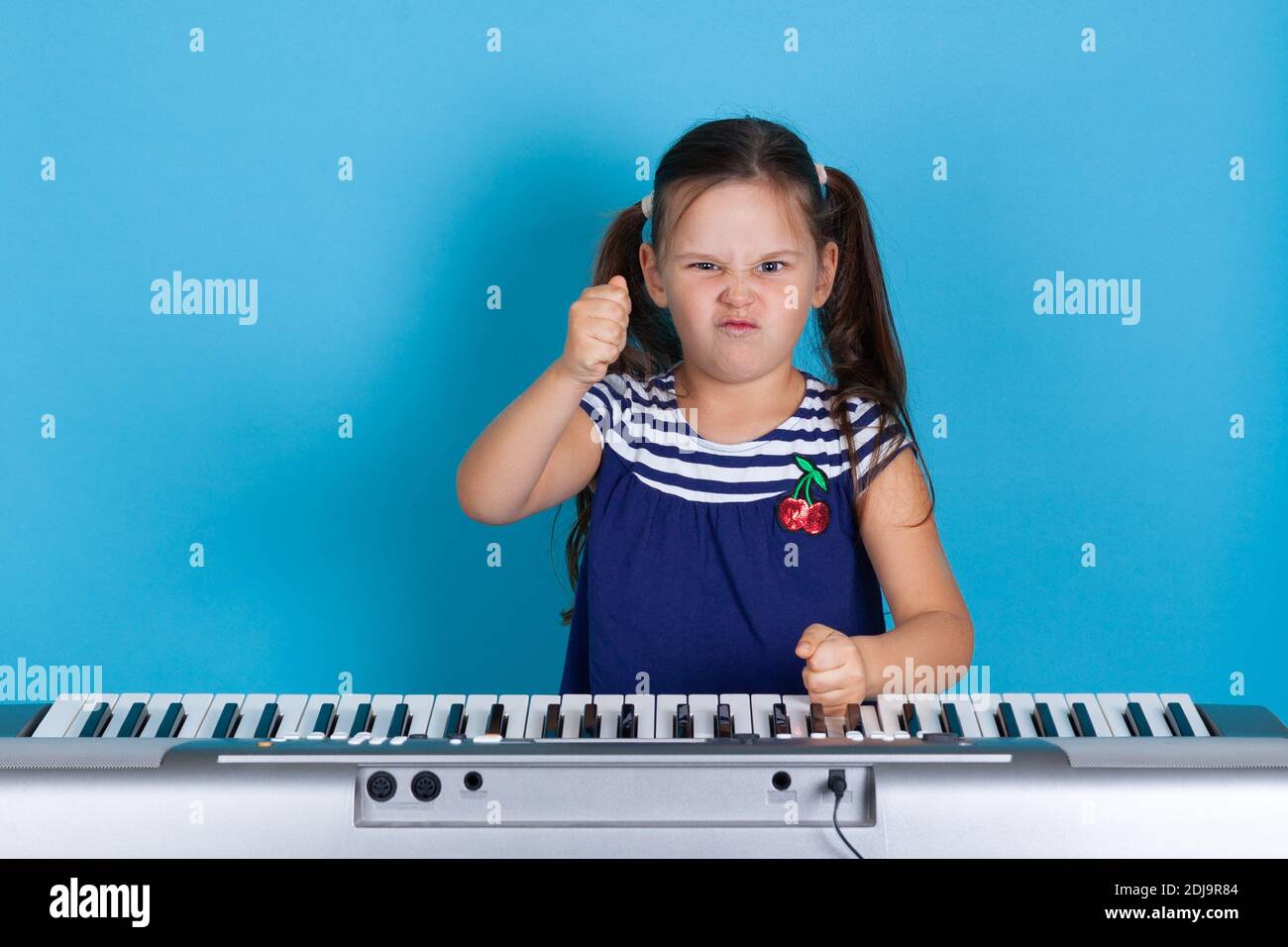 primo piano di una ragazza malvagia che batte i pugni sui tasti del sintetizzatore, non gradito per le classi di musica online, isolato su uno sfondo blu Foto Stock