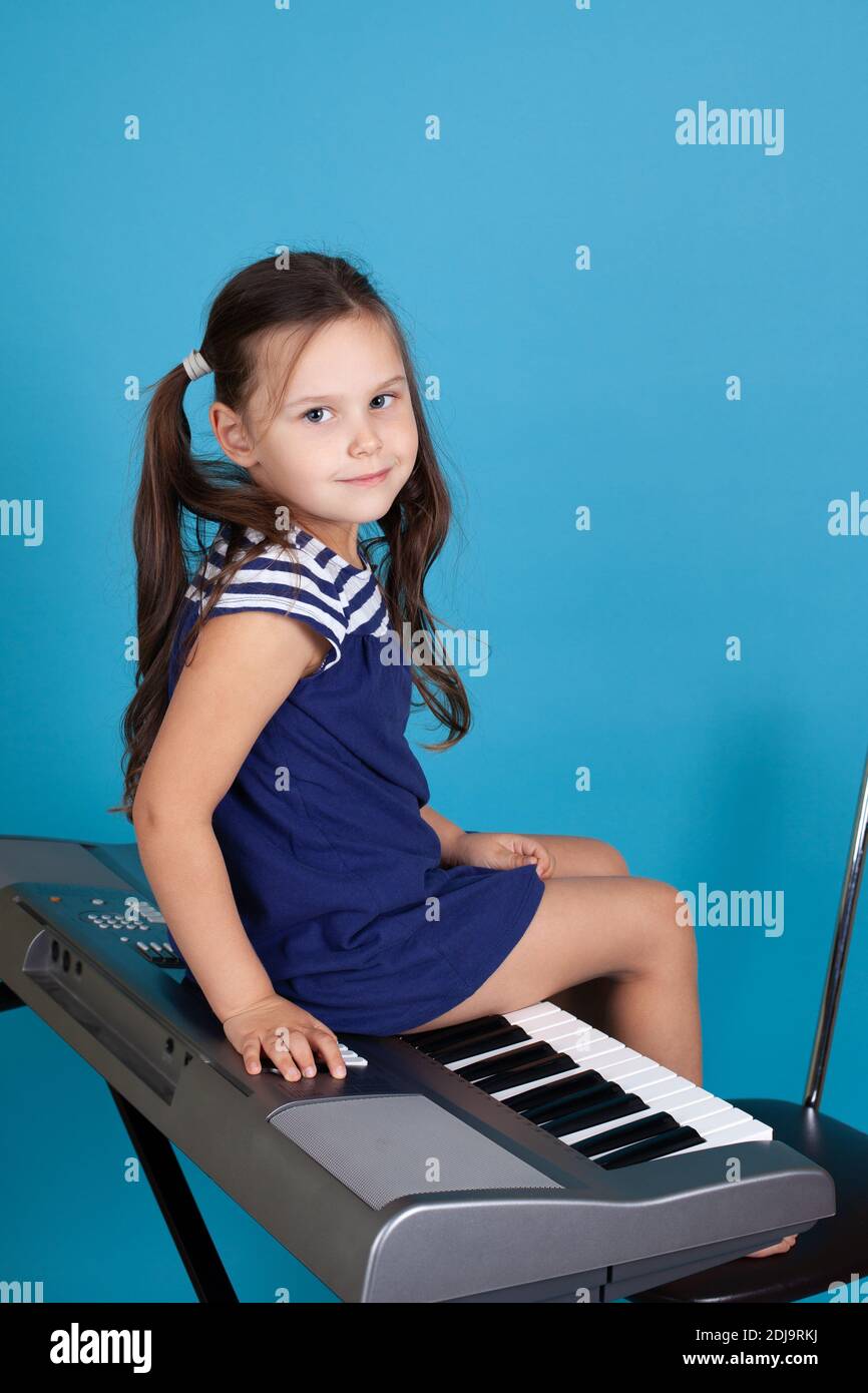una ragazza sorridente di 5 anni si siede a cavallo di un sintetizzatore elettronico, isolato su uno sfondo blu Foto Stock
