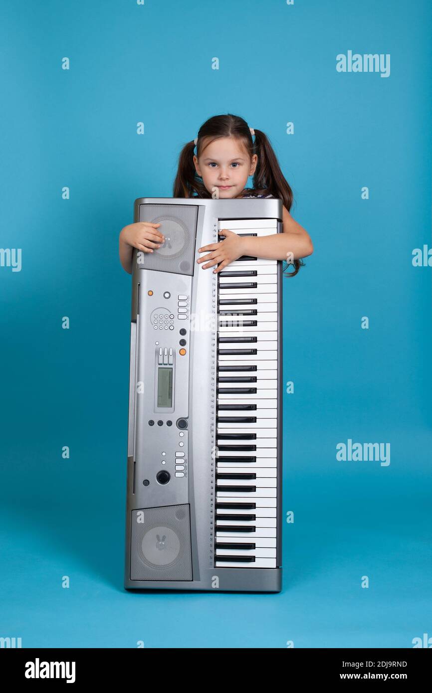 ritratto a tutta lunghezza di un bel bambino che abbraccia un sintetizzatore elettronico verticale e si nasconde dietro di esso, isolato su sfondo blu Foto Stock