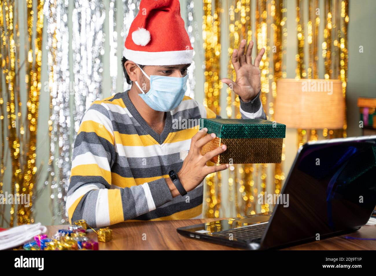 Giovane uomo in maschera di faccia che mostra la scatola del regalo durante in linea video chat su laptop durante il nuovo anno o le festività natalizie celebrazione Foto Stock