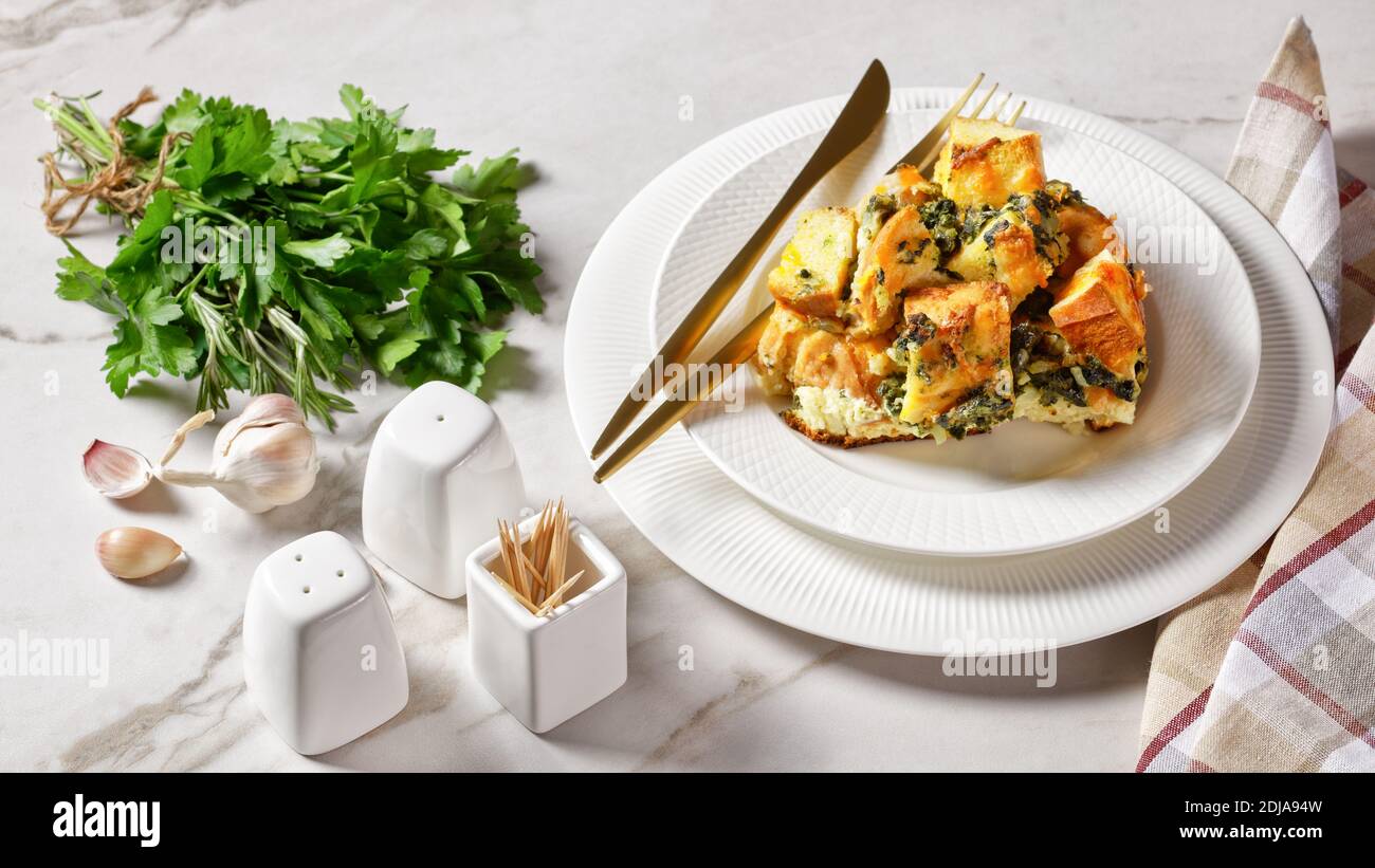 Strata o casseruola italiana per la colazione di spinaci, formaggio e pane ammollito per una notte al forno con senape su un piatto bianco con posate dorate su a. Foto Stock
