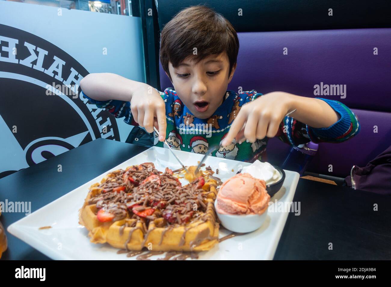 Un ragazzo che indossa un jumper natalizio mangia un grande waffle con gelato, fragole e cioccolato. Foto Stock