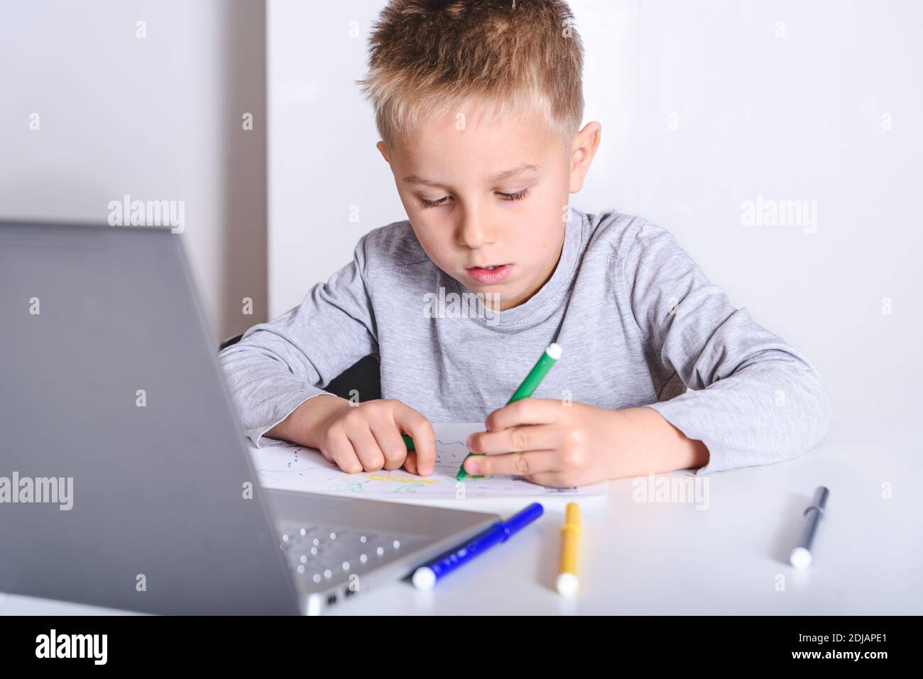 biondo caucasico ragazzino di 5 anni disegno con feltro penne in soggiorno a casa su un tavolo bianco con un computer portatile e con sfondo bianco Foto Stock