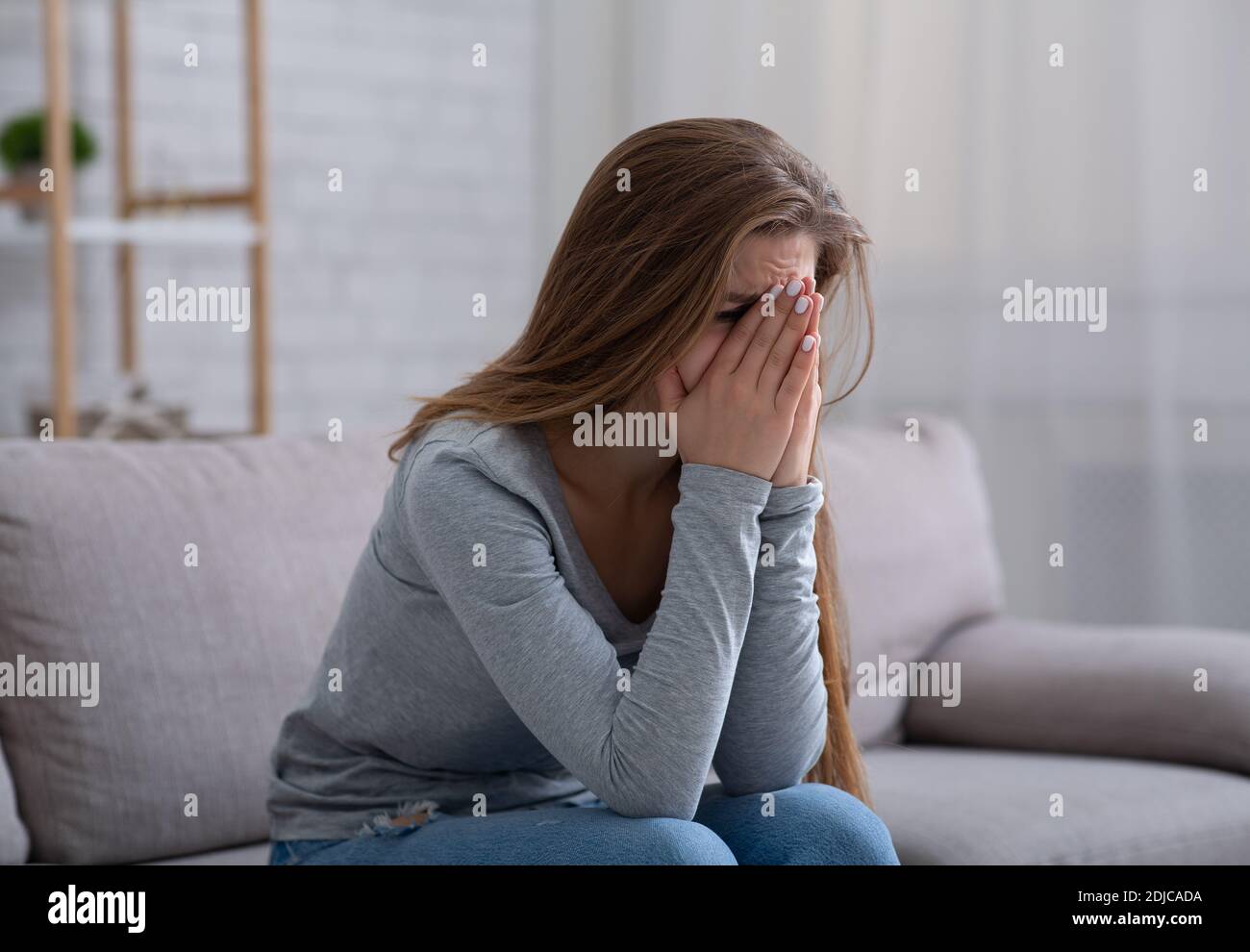 Giovane donna che seppellisce il viso in mani e piangendo, sentendosi depresso o disperato sul divano al chiuso Foto Stock