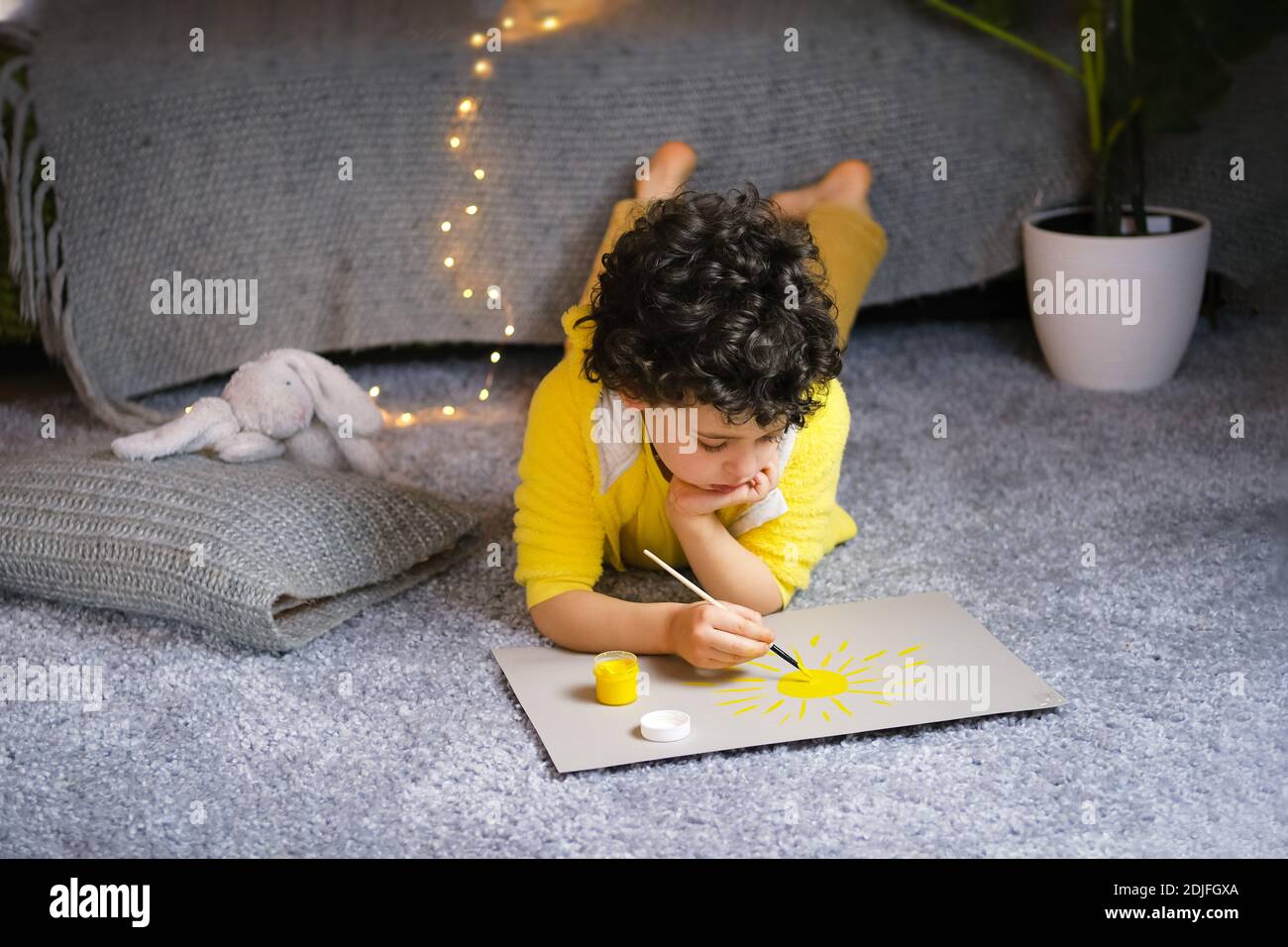 Piccolo ragazzo riccamente disegnando il sole giallo in interni grigi a casa. Foto Stock
