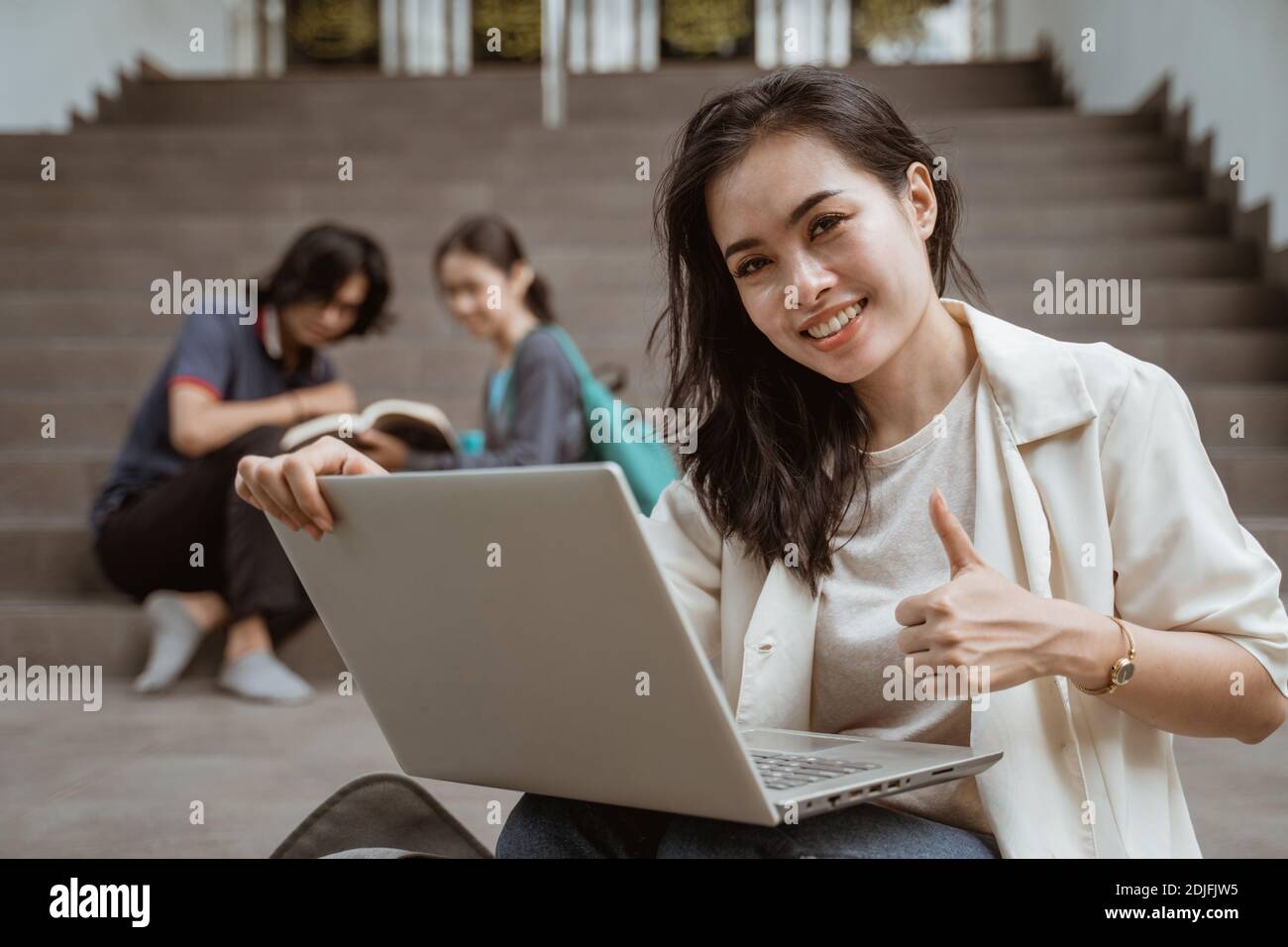 Ritratti felici gli studenti che tengono un computer portatile e mostrano i pollici con gli amici sullo sfondo Foto Stock