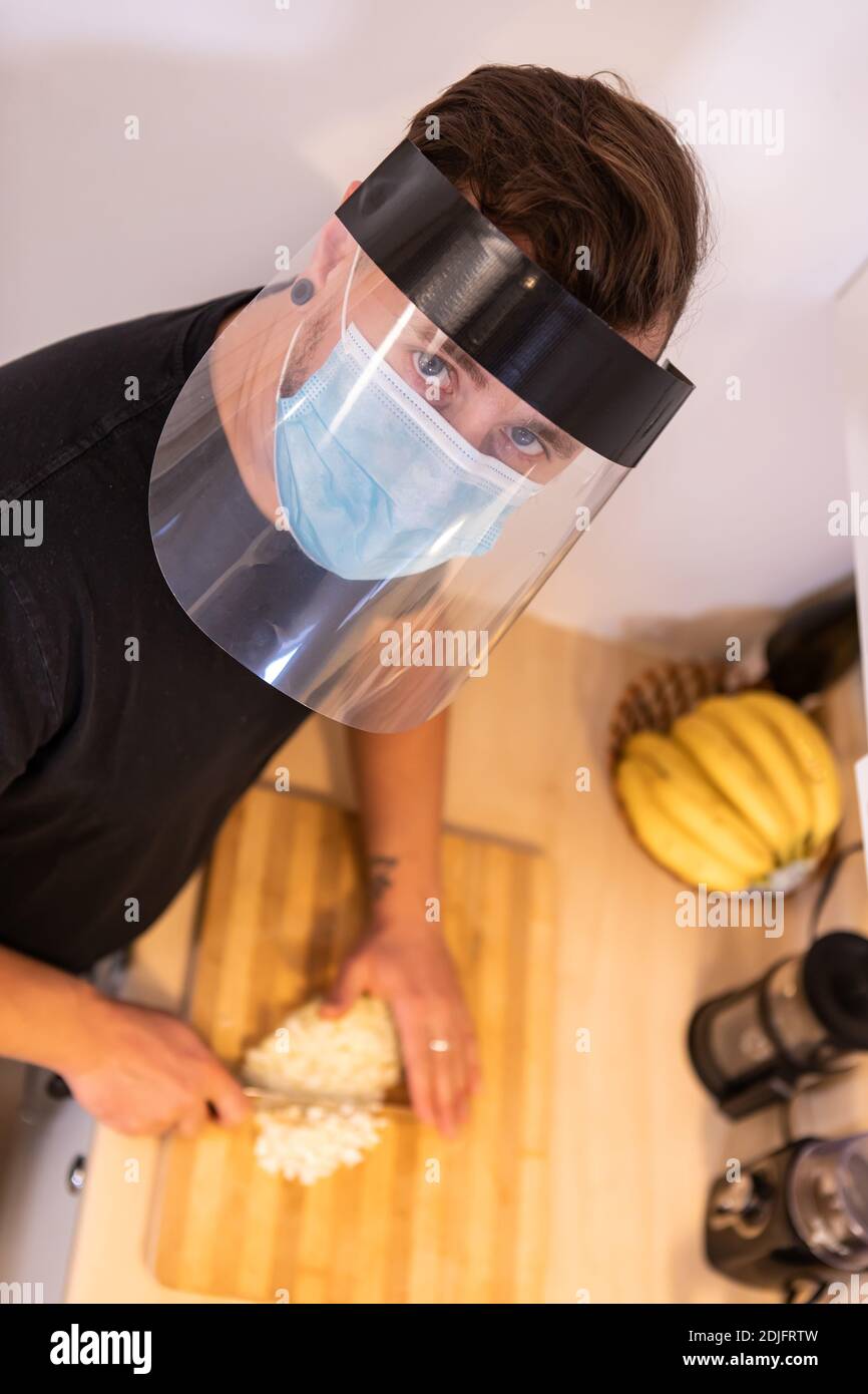 Primo piano dall'alto di un giovane uomo, guardando la macchina fotografica e tagliando il cibo su un cinghiale da taglio con maschera protettiva e visiera in plastica Foto Stock