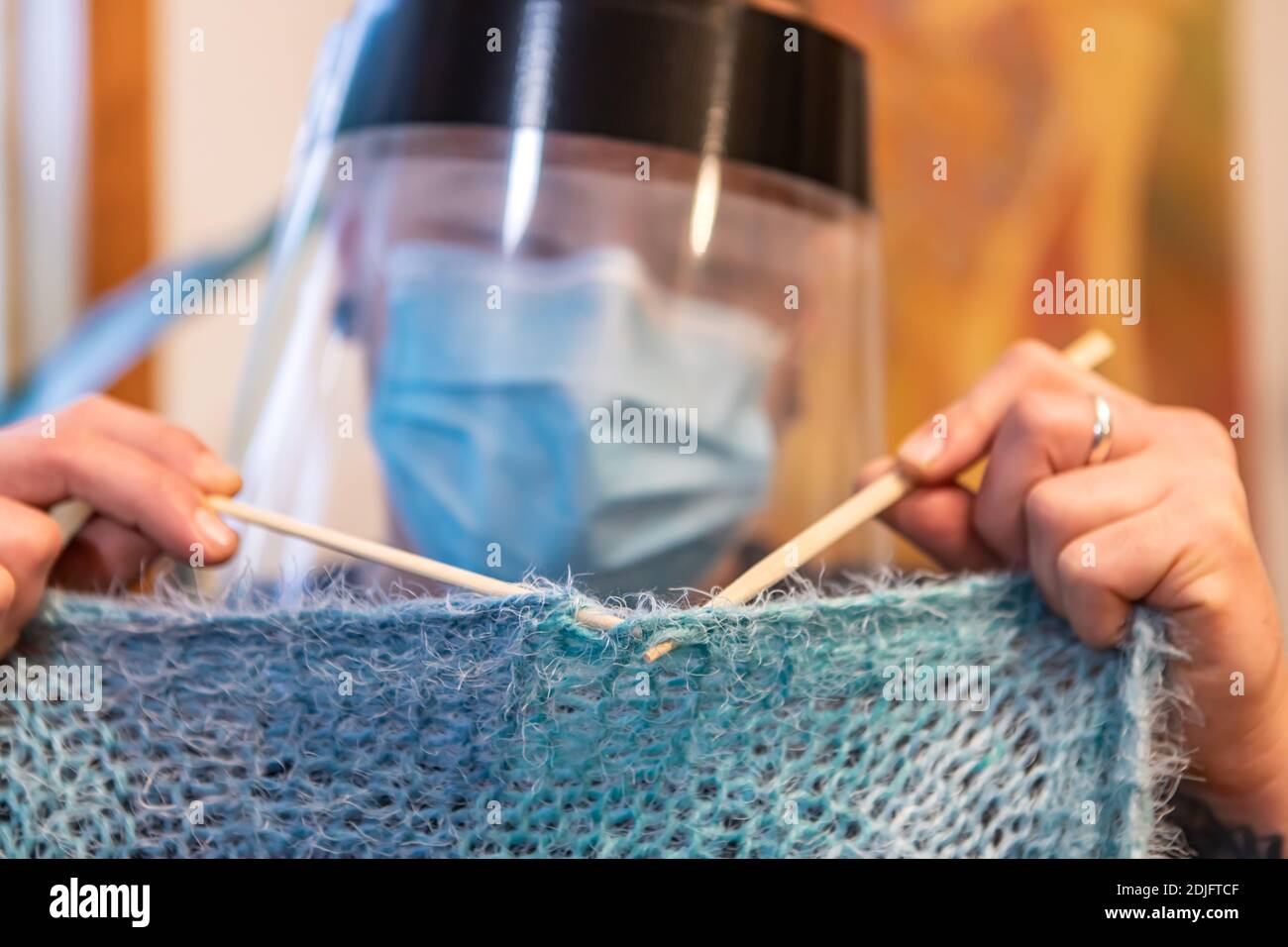 Divertente primo piano di un giovane uomo indoor, lana di maglieria con aghi da maglieria, mentre si indossa una maschera di protezione covid e visiera in plastica. Foto Stock