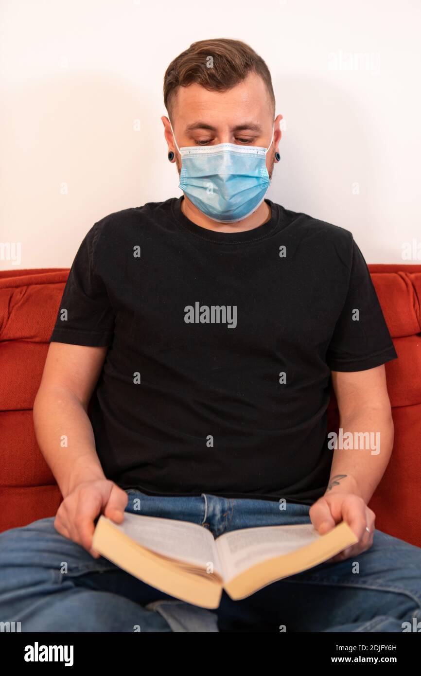 Stile di vita Covid. Eccessiva prevenzione del covidio. Uomo seduto a croce zampe su un divano rosso con un libro che indossa una maschera sanitaria contro covid. Scatto ironico. Foto Stock