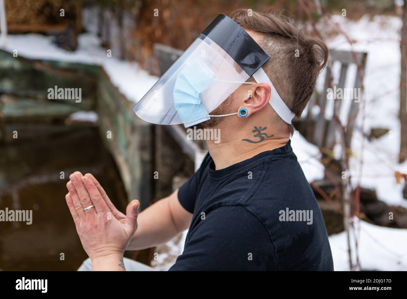 Vista laterale di un uomo in una t-shirt seduta sul bordo o fa lago nella neve, indossando maschera covid e visiera protettiva e unendosi le mani in preghiera. Foto Stock