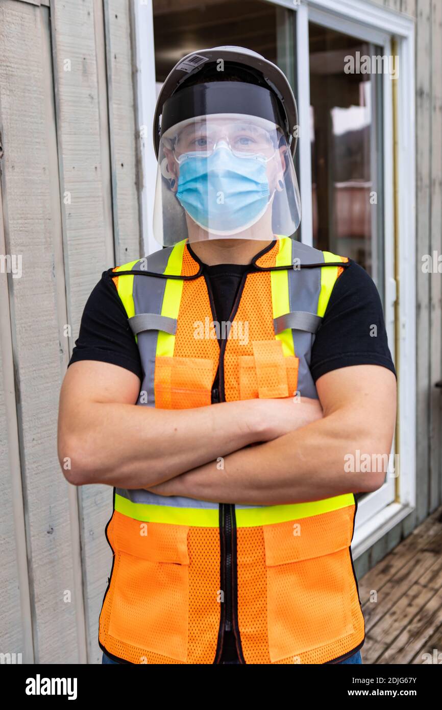 colpo medio vicino di un uomo in piedi braccia incrociate indossare una giacca arancione e giallo fluo visibilità, covid maschera protettiva viso e visiera in plastica. Foto Stock