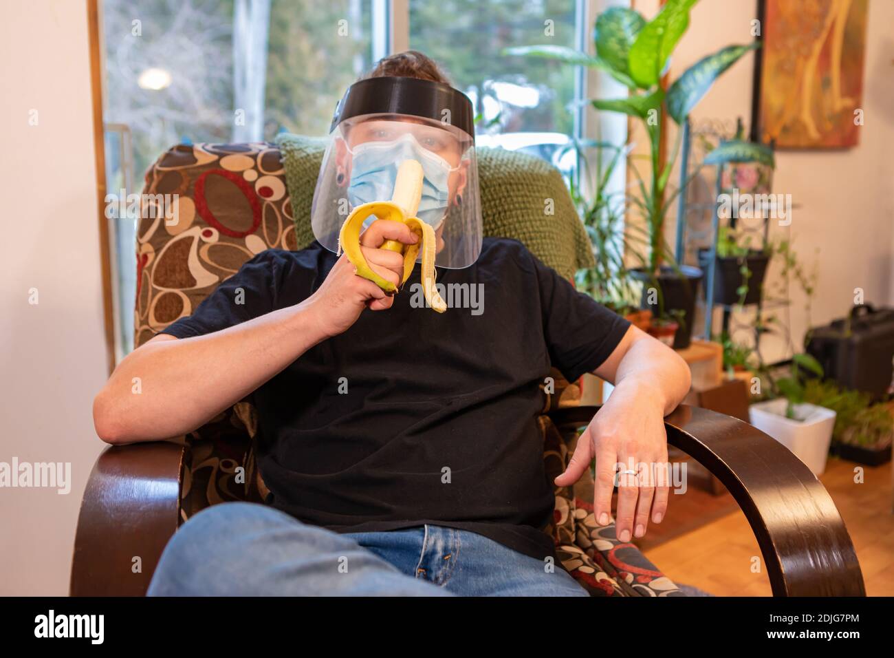 Scatto medio vicino di un giovane uomo che si rilassa in una poltrona interna e mangia una banana, indossando maschera protettiva e visiera in plastica anticovida. Foto Stock