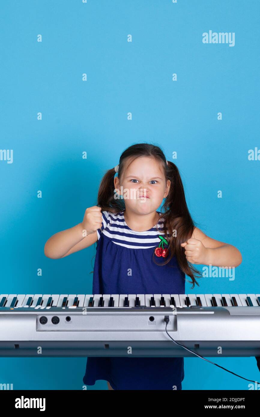 vista frontale di una ragazza accigliata che batte i pugni sui tasti del sintetizzatore, arrabbiata a una lezione di musica isolata su uno sfondo blu Foto Stock