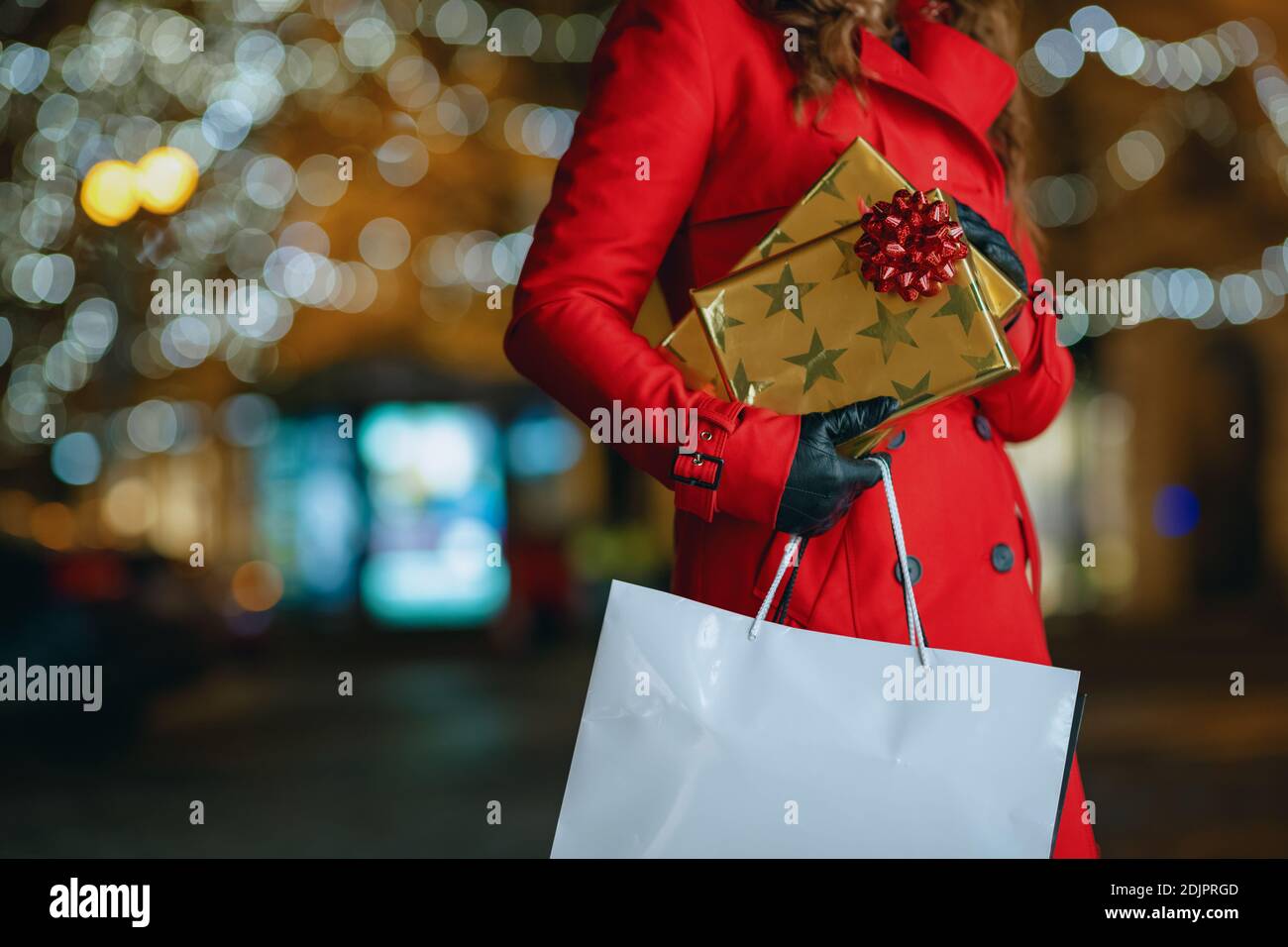 Divertimento invernale. Primo piano su donna viaggiatore di mezza età in berretto rosso e nero con borse per lo shopping e regali all'aperto in città in serata. Foto Stock