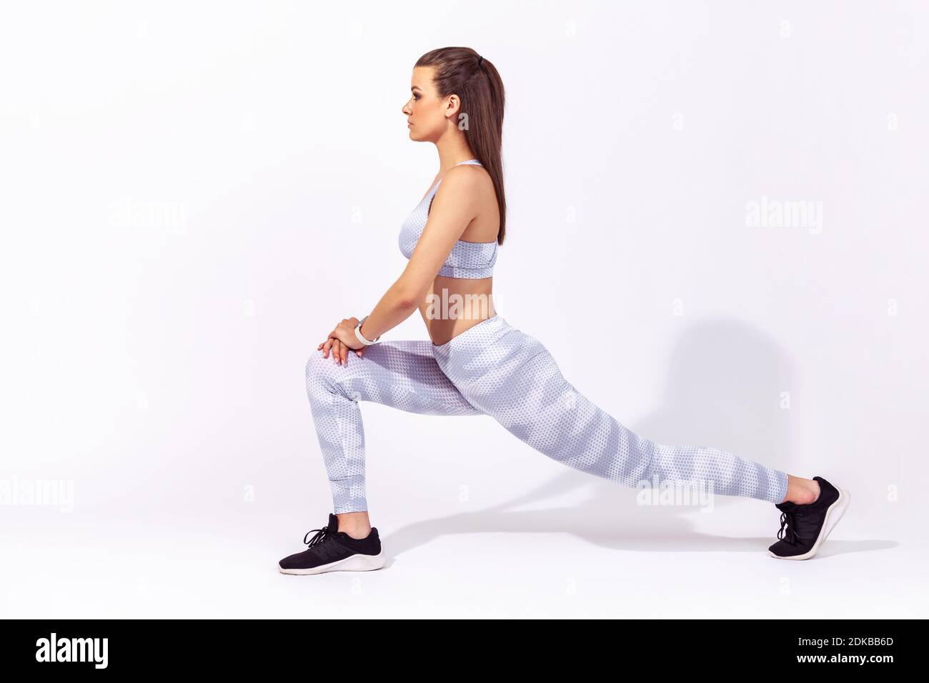 Profilo ritratto full length donna atletica in bianco sportswear facendo esercizio sportivo corpo inferiore, allungando le gambe, riscaldando i muscoli per la flessibilità. Foto Stock
