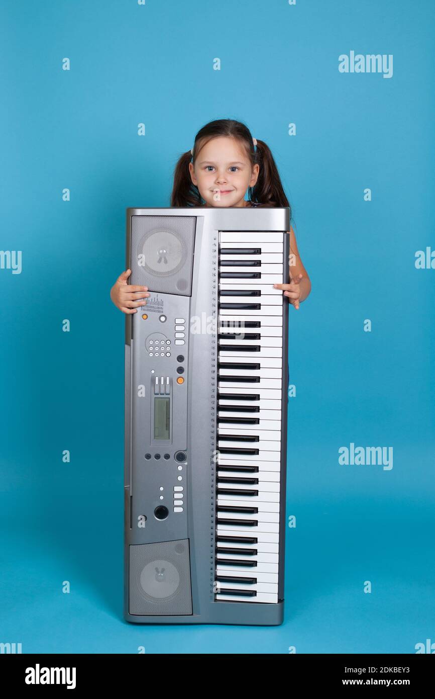 ritratto a tutta lunghezza di una ragazza sorridente che si nasconde dietro un sintetizzatore elettronico verticale e suona i suoi tasti, isolato su uno sfondo blu Foto Stock