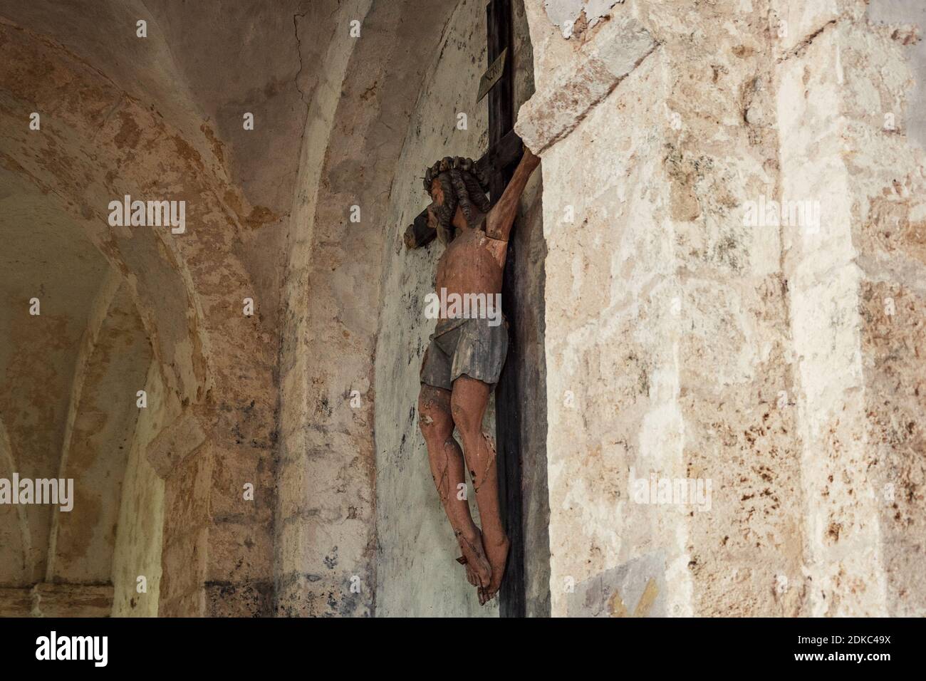 Vecchio crocifisso di legno con peeling vernice contro grugnoso muro di pietra sporco sfondo in chiesa. Concetto di problema di conservazione del patrimonio religioso. Foto Stock