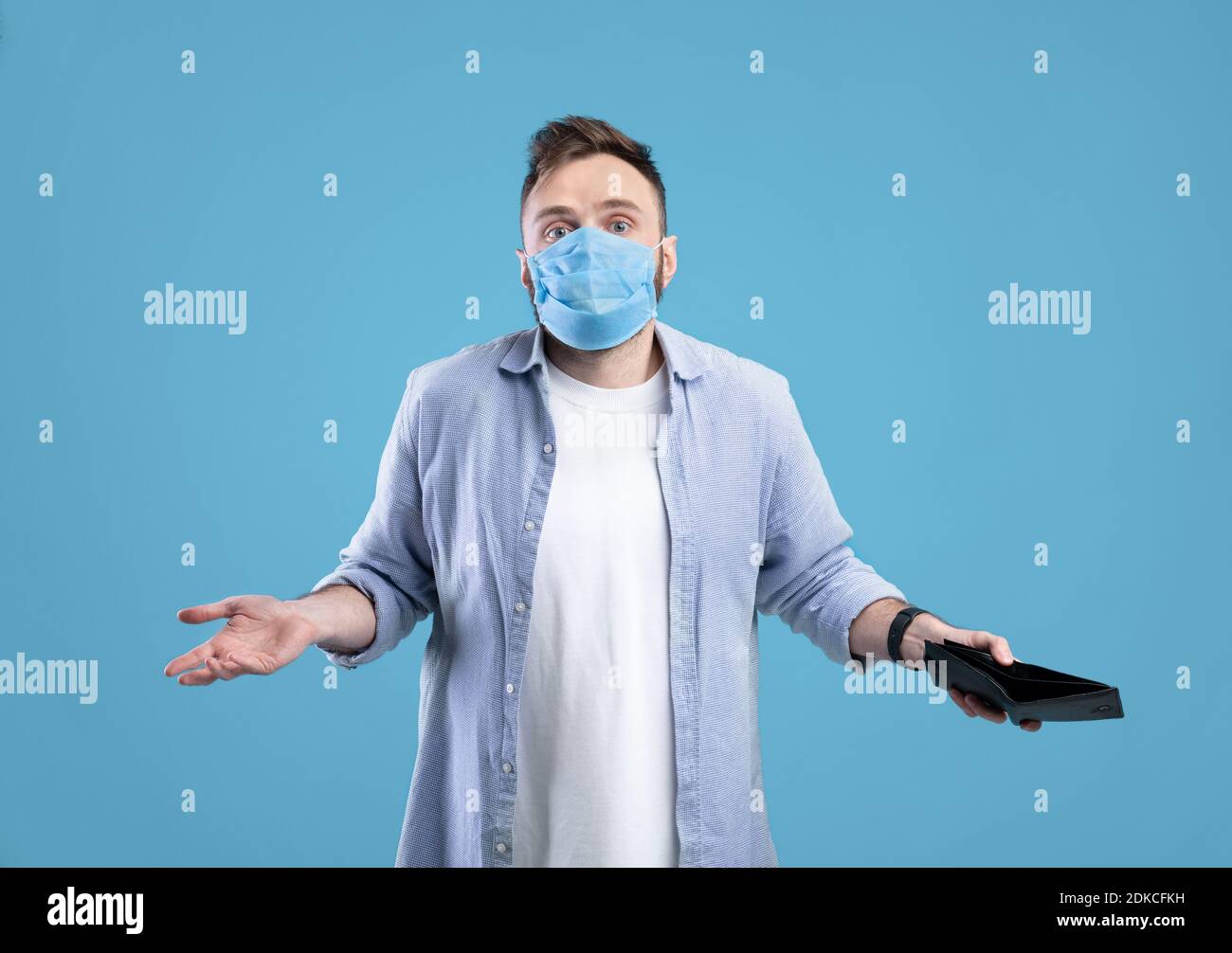 Giovane uomo in maschera protettiva che tiene il portafoglio vuoto con no soldi e spalle di rugging su sfondo blu studio Foto Stock